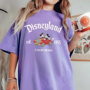 K&ouml;nnte beinhalten: Ein lila T-Shirt mit dem Disneyland-Logo in wei&szlig;er Schrift und "est. 1955 CALIFORNIA" darunter. Auf dem T-Shirt befindet sich eine Grafik von Mickey und Minnie Mouse in einem roten Auto. Das T-Shirt hat kurze &Auml;rmel.
