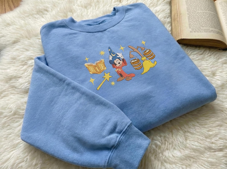 May include: A pale blue sweatshirt with an embroidered cartoon design of a wizard character, a book, a wand, and a broom. The sweatshirt has a crew neck and long sleeves. The design is in shades of yellow, red, and blue.