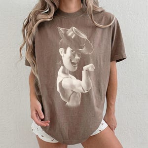 Puede incluir: Una camiseta marr&oacute;n de manga corta con un gran estampado gr&aacute;fico monocrom&aacute;tico de un personaje vaquero con sombrero de ala ancha. El personaje aparece desde el pecho hacia arriba, sonriendo con un brazo doblado por el codo.