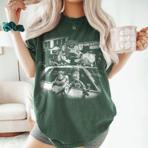 May include: A dark green short-sleeve t-shirt featuring a distressed white graphic print of characters from the Toy Story franchise depicted in a gritty, cinematic style. The design shows multiple characters positioned around a car, with some holding firearms.