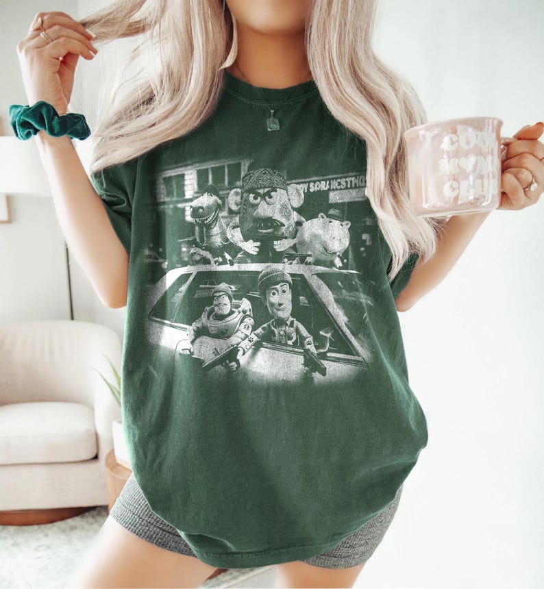 May include: A dark green short-sleeve t-shirt featuring a distressed white graphic print of characters from the Toy Story franchise depicted in a gritty, cinematic style. The design shows multiple characters positioned around a car, with some holding firearms.