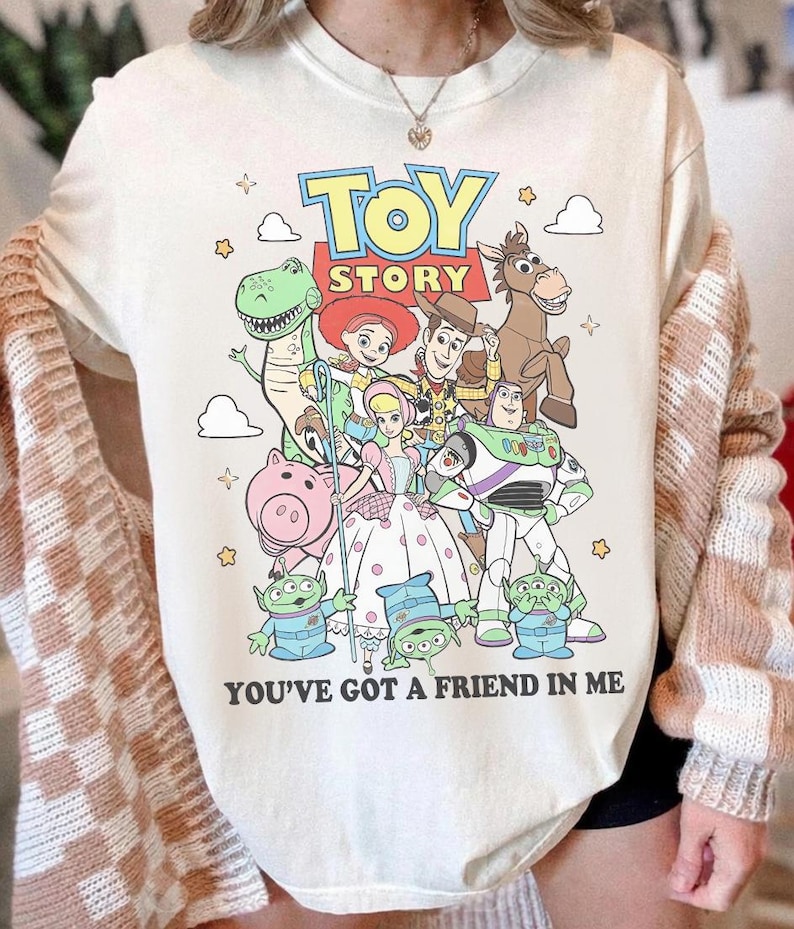 May include: A cream-colored t-shirt featuring a colorful graphic of Toy Story characters including Woody, Buzz Lightyear, Jessie, Rex, Bullseye, Hamm, Bo Peep, and the Aliens. The text reads TOY STORY and YOU'VE GOT A FRIEND IN ME with small stars.