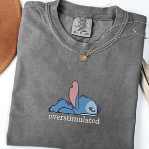 May include: A charcoal gray t-shirt featuring an embroidered graphic of the blue character Stitch lying down with the text "overstimulated" printed in white underneath. The shirt has a Comfort Colors brand label at the inner neckline.