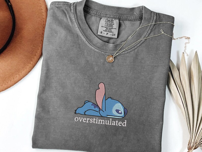 May include: A charcoal gray t-shirt featuring an embroidered graphic of the blue character Stitch lying down with the text "overstimulated" printed in white underneath. The shirt has a Comfort Colors brand label at the inner neckline.