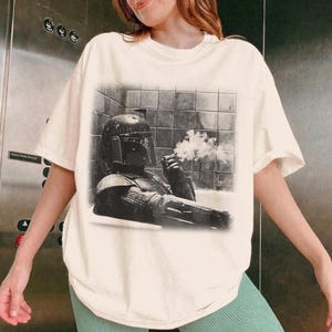 May include: An off-white short-sleeve t-shirt featuring a black and white graphic print of a helmeted character in a bathtub, holding a cigar with visible smoke. The graphic shows the character wearing armor and a helmet while sitting in a tiled tub.