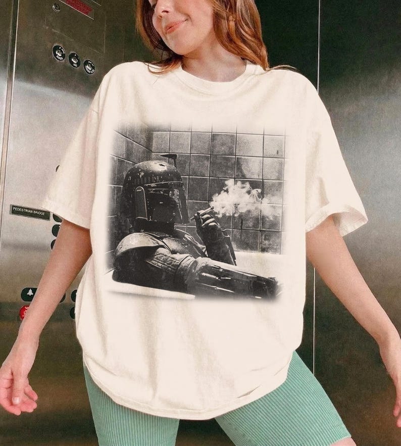 May include: An off-white short-sleeve t-shirt featuring a black and white graphic print of a helmeted character in a bathtub, holding a cigar with visible smoke. The graphic shows the character wearing armor and a helmet while sitting in a tiled tub.