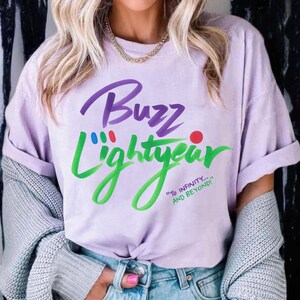 Puede incluir: Una camiseta de manga corta de color morado claro con el texto Buzz Lightyear en letra cursiva morada y verde. El dise&ntilde;o incluye peque&ntilde;os puntos rojos, azules y verdes sobre el texto y la frase To Infinity... And Beyond! en verde.