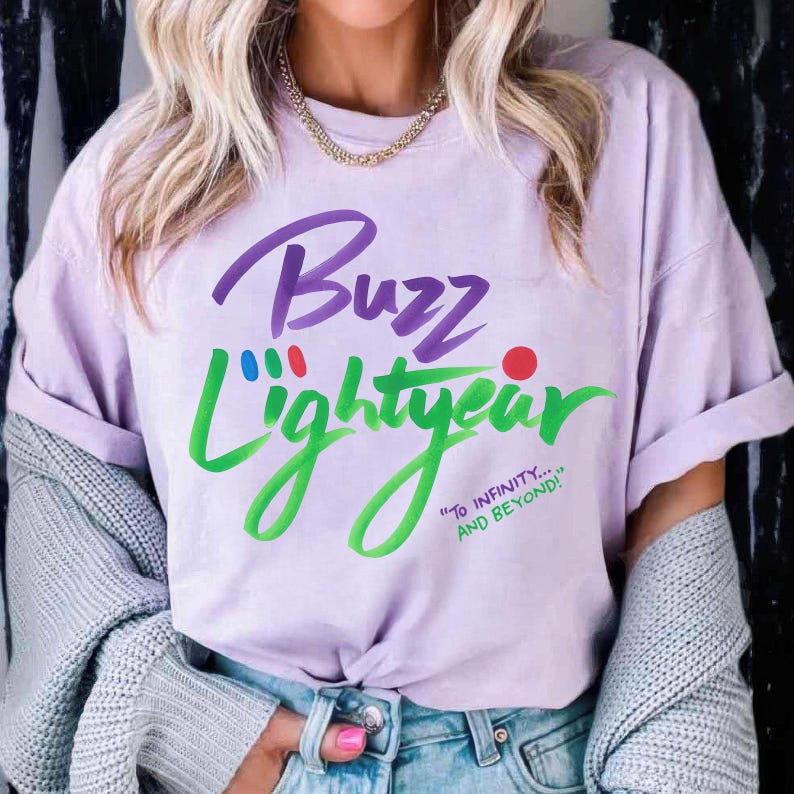 Puede incluir: Una camiseta de manga corta de color morado claro con el texto Buzz Lightyear en letra cursiva morada y verde. El dise&ntilde;o incluye peque&ntilde;os puntos rojos, azules y verdes sobre el texto y la frase To Infinity... And Beyond! en verde.