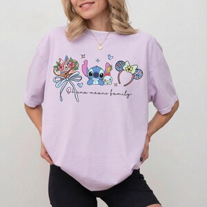 Puede incluir: Camiseta lila claro con un gr&aacute;fico colorido de Stitch, detalles florales y la frase "Ohana means family". El dise&ntilde;o incluye un lazo, estrellas y una diadema con detalles florales. La camiseta es de estilo c&oacute;modo e informal.