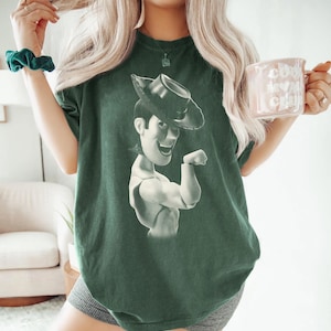 Puede incluir: Una camiseta de manga corta de color verde oscuro con un estampado gr&aacute;fico monocrom&aacute;tico del personaje Woody de Toy Story con un sombrero de vaquero. La prenda tiene cuello redondo y un corte holgado, mostrando al personaje de perfil.