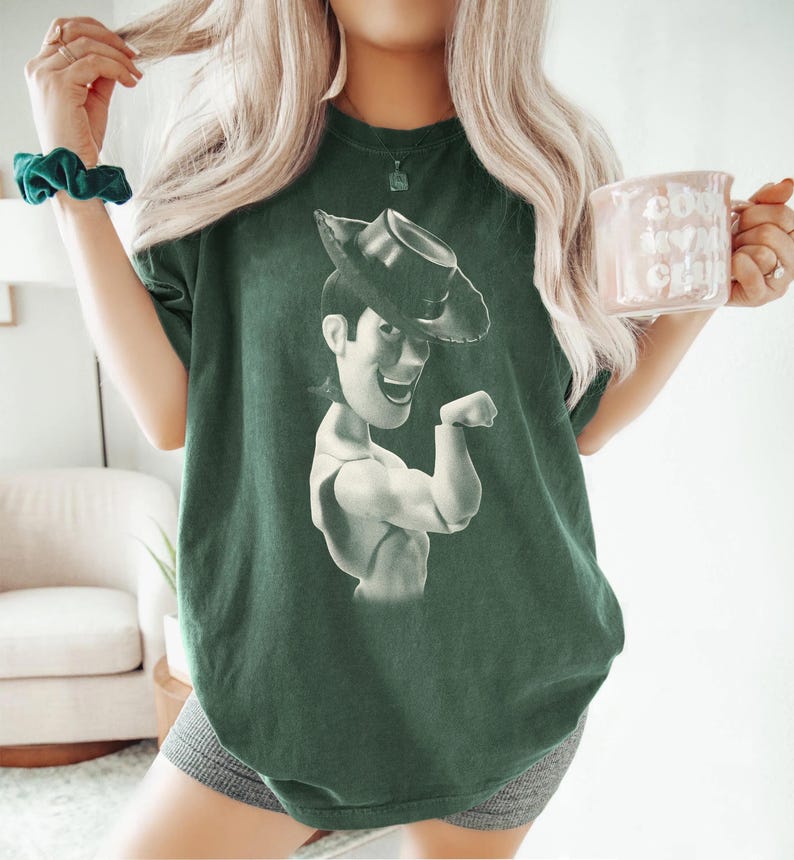 Puede incluir: Una camiseta de manga corta de color verde oscuro con un estampado gr&aacute;fico monocrom&aacute;tico del personaje Woody de Toy Story con un sombrero de vaquero. La prenda tiene cuello redondo y un corte holgado, mostrando al personaje de perfil.