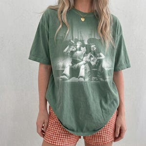 May include: A sage green short-sleeve t-shirt featuring a monochromatic graphic print of two characters sitting together. The shirt has a crew neckline and a relaxed fit. The graphic depicts a space ranger figure and a cowboy figure in a dark setting.