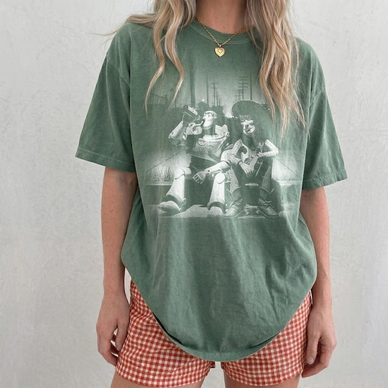 May include: A sage green short-sleeve t-shirt featuring a monochromatic graphic print of two characters sitting together. The shirt has a crew neckline and a relaxed fit. The graphic depicts a space ranger figure and a cowboy figure in a dark setting.