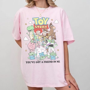 May include: A light pink t-shirt featuring a colorful graphic of Toy Story characters including Woody, Buzz Lightyear, Jessie, Rex, Bullseye, Hamm, Bo Peep, and the Aliens. The text TOY STORY and YOU'VE GOT A FRIEND IN ME are printed on the front.