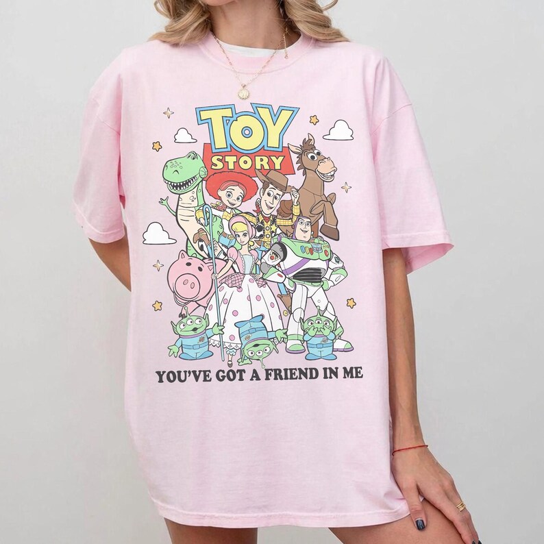 May include: A light pink t-shirt featuring a colorful graphic of Toy Story characters including Woody, Buzz Lightyear, Jessie, Rex, Bullseye, Hamm, Bo Peep, and the Aliens. The text TOY STORY and YOU'VE GOT A FRIEND IN ME are printed on the front.