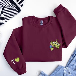 May include: A maroon long-sleeve sweatshirt featuring an embroidered green three-eyed alien character on the chest. The left sleeve displays the white letter T followed by a green embroidered heart. The garment has a crew neckline and ribbed cuffs.