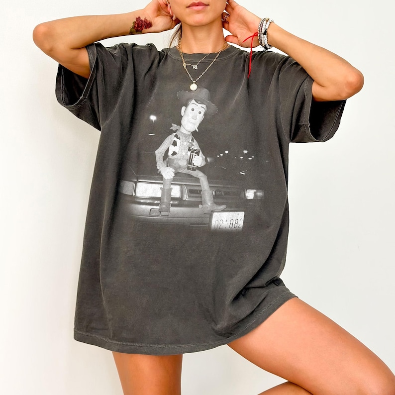 May include: A charcoal gray short-sleeve t-shirt featuring a black and white graphic print of a cowboy character sitting on the hood of a car. The graphic includes a license plate with the text 02-8820 visible on the front bumper of the vehicle.