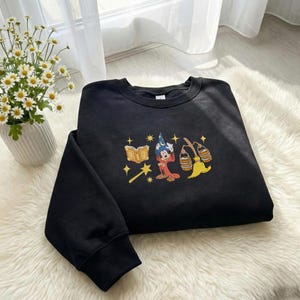 May include: Black crew neck sweatshirt with an embroidered design of a cartoon wizard character, a book, a magic wand, and a broom. The design is in red, yellow, blue, and gold. The sweatshirt is on a white fluffy surface.