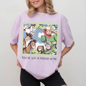 May include: A light purple t-shirt featuring a colorful graphic of characters from the animated movie Toy Story. The graphic is framed with a green border and includes the text "YOU'VE GOT A FRIEND IN ME".