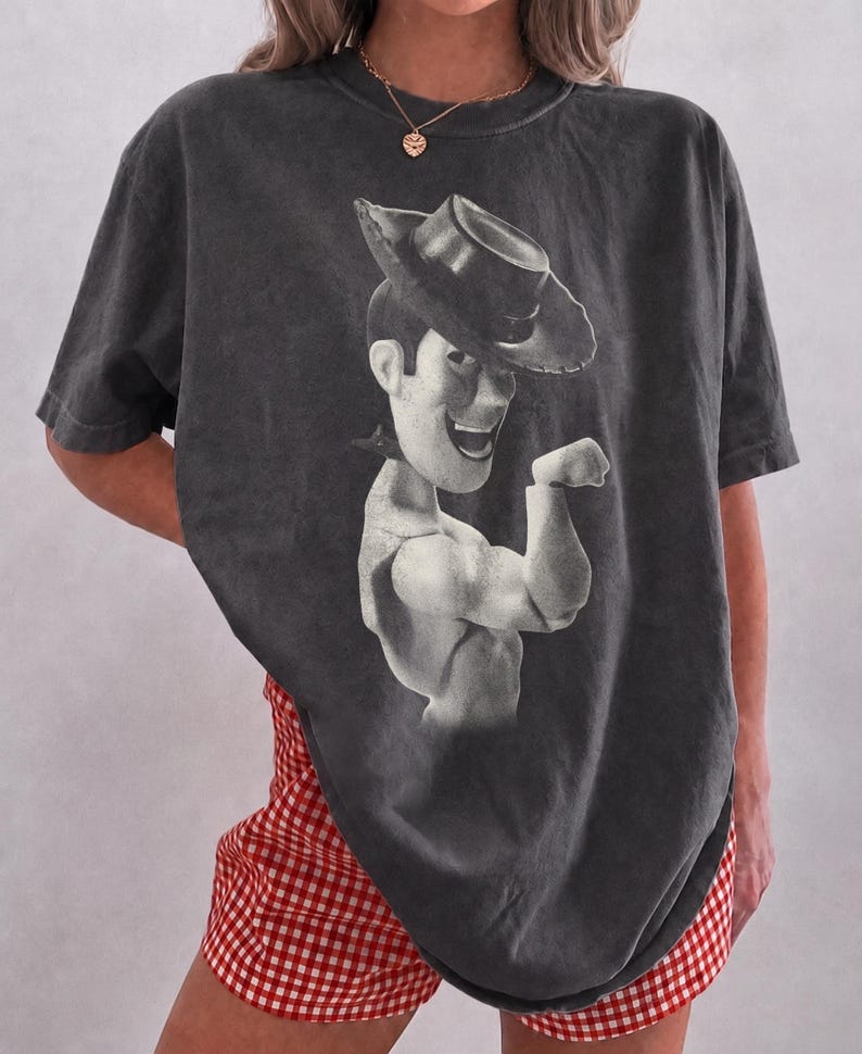 Puede incluir: Una camiseta de manga corta en color gris carb&oacute;n con un gran estampado gr&aacute;fico monocrom&aacute;tico del personaje Woody de Toy Story con sombrero de vaquero. La camiseta tiene un corte holgado y extragrande, combinada con pantalones cortos de cuadros vichy rojos y blancos.
