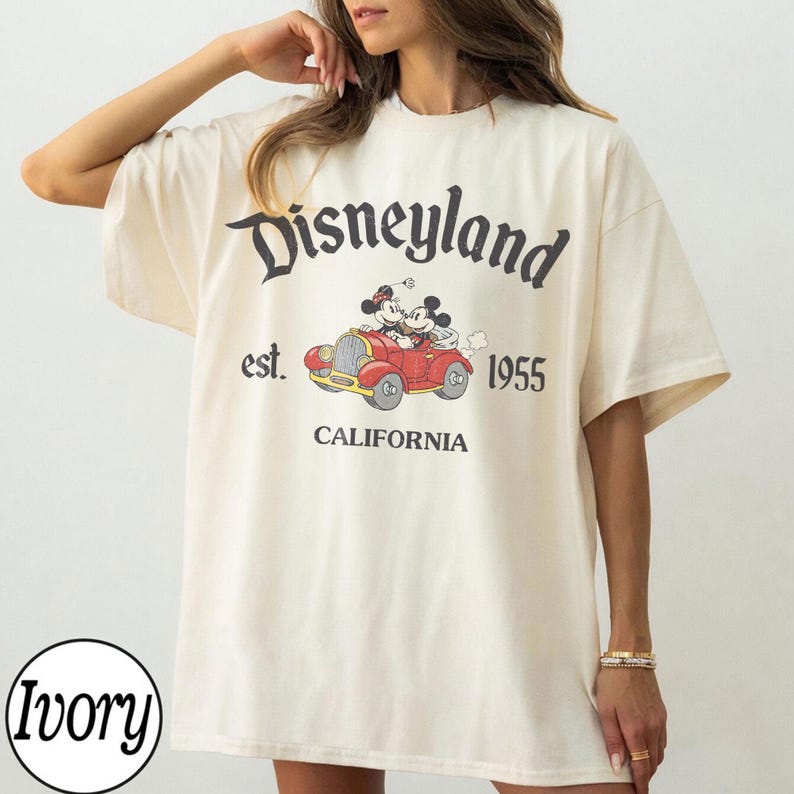 K&ouml;nnte beinhalten: Elfenbeinfarbenes T-Shirt mit dem Disneyland-Logo und einer Grafik von Mickey und Minnie Mouse in einem roten Auto. Der Text "est. 1955 CALIFORNIA" ist in Schwarz aufgedruckt. Das T-Shirt hat kurze &Auml;rmel und eine lockere Passform.