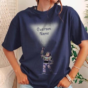 Pu&ograve; includere: T-shirt blu navy con una grafica di Buzz Lightyear di Toy Story e la scritta "Custom Name" in una nuvola bianca. L'astronauta dei cartoni animati &egrave; raffigurato in una posa dinamica, con il braccio alzato.