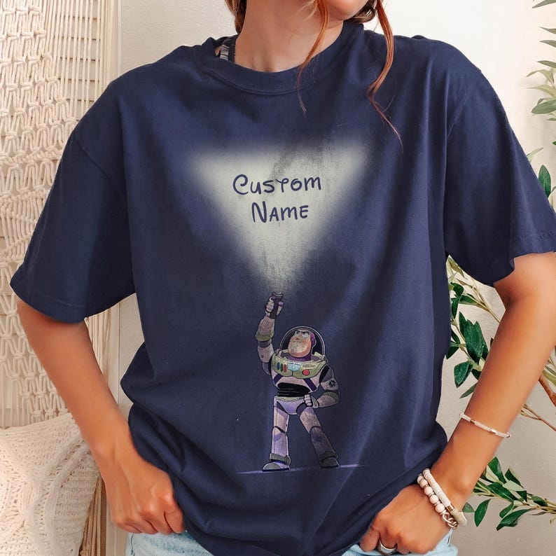 Pu&ograve; includere: T-shirt blu navy con una grafica di Buzz Lightyear di Toy Story e la scritta "Custom Name" in una nuvola bianca. L'astronauta dei cartoni animati &egrave; raffigurato in una posa dinamica, con il braccio alzato.