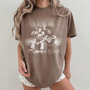 May include: A brown short-sleeve t-shirt featuring a monochromatic graphic print of two animated characters sitting side-by-side. The design depicts a space ranger figure and a cowboy figure in a faded, distressed style on the front of the shirt.