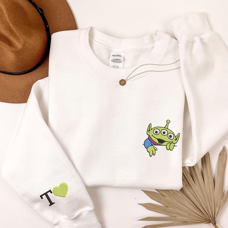 May include: A white long-sleeve crewneck sweatshirt featuring an embroidered green three-eyed alien character on the chest. The left sleeve displays an embroidered black letter T followed by a small green heart symbol, creating a simple design.