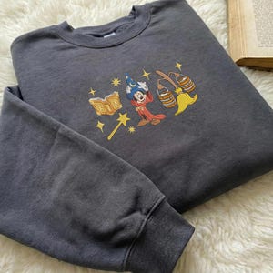 May include: Dark grey sweatshirt with an embroidered design of a cartoon wizard character, a book, a magic wand, and brooms. The design uses red, yellow, blue, and gold. The sweatshirt has a crew neck and long sleeves.