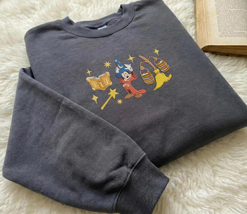 May include: Dark grey sweatshirt with an embroidered design of a cartoon wizard character, a book, a magic wand, and brooms. The design uses red, yellow, blue, and gold. The sweatshirt has a crew neck and long sleeves.