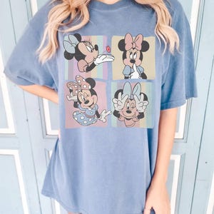 May include: Blue t-shirt featuring a four-panel graphic of Minnie Mouse in various poses. Each panel has a different background color and a different Minnie Mouse design. The shirt is a solid blue color.