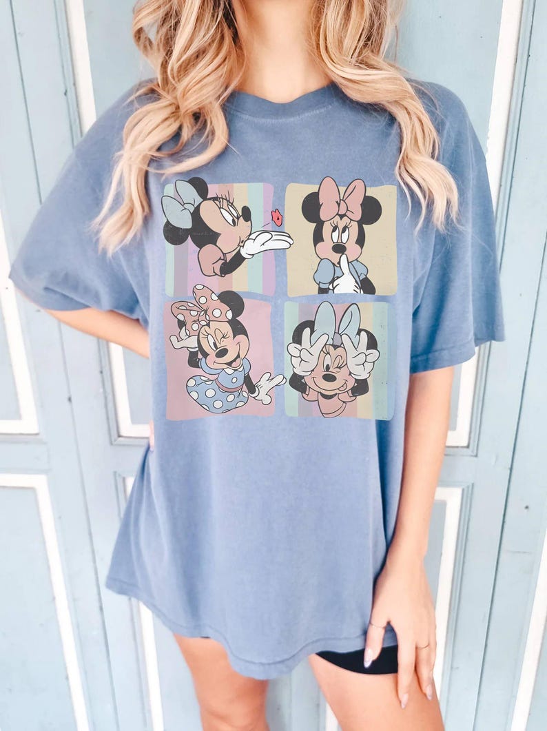 May include: Blue t-shirt featuring a four-panel graphic of Minnie Mouse in various poses. Each panel has a different background color and a different Minnie Mouse design. The shirt is a solid blue color.