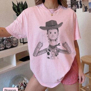 May include: A light pink short-sleeve t-shirt featuring a black and white graphic print of a cowboy character wearing a hat, a vest, and a neckerchief, with hands positioned near the face and tongue sticking out in a playful expression.