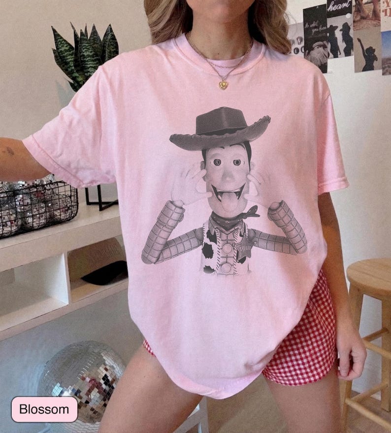 May include: A light pink short-sleeve t-shirt featuring a black and white graphic print of a cowboy character wearing a hat, a vest, and a neckerchief, with hands positioned near the face and tongue sticking out in a playful expression.