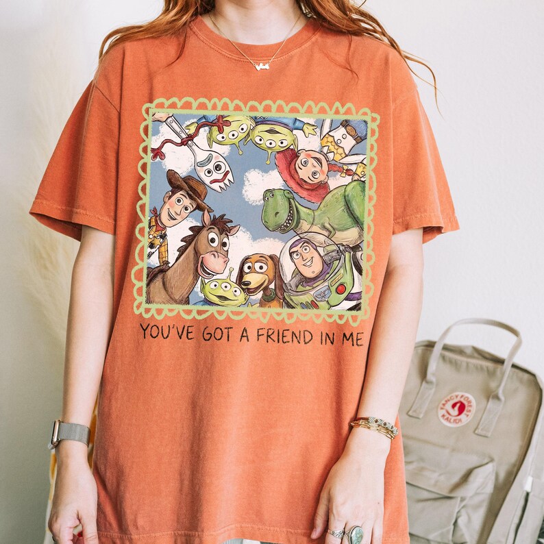 May include: Orange t-shirt featuring a framed illustration of characters from the animated film "Toy Story." The image includes Woody, Buzz Lightyear, Rex, and other characters. The text "YOU'VE GOT A FRIEND IN ME" is printed below the illustration.