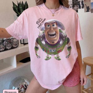 May include: A light pink short-sleeve t-shirt featuring a graphic of the character Buzz Lightyear in a space suit with a clear helmet. The text "BUZZ LIGHTYEAR" is printed in black script above the character. The shirt has a crew neckline.