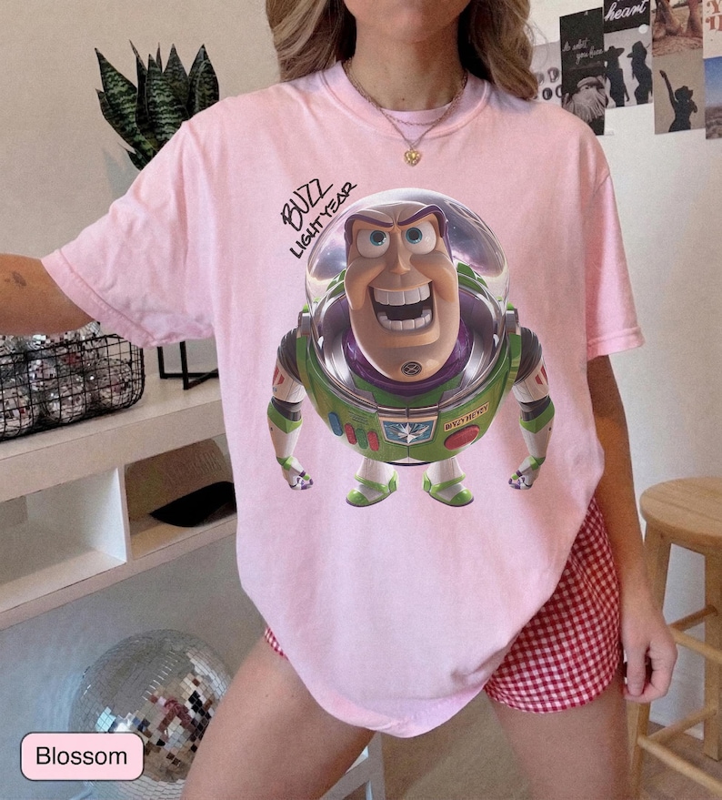 May include: A light pink short-sleeve t-shirt featuring a graphic of the character Buzz Lightyear in a space suit with a clear helmet. The text "BUZZ LIGHTYEAR" is printed in black script above the character. The shirt has a crew neckline.