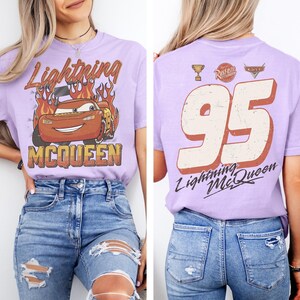 May include: Lavender t-shirt featuring a graphic of Lightning McQueen from the Cars movie, with flames and the text "Lightning McQueen" and "MCQUEEN". The back of the shirt displays the number "95" and the text "Lightning McQueen".