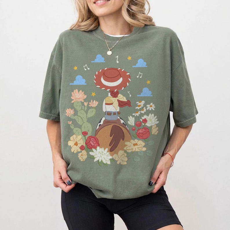 May include: An olive green short-sleeve t-shirt featuring a graphic print of a cowgirl figure wearing a red hat, riding a horse, surrounded by colorful flowers, cacti, blue clouds, and yellow stars against a muted, earthy background.