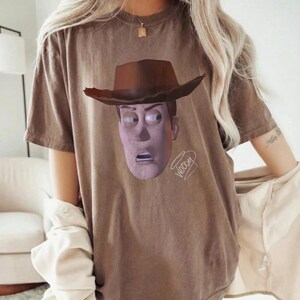 May include: A brown short-sleeved t-shirt featuring a graphic print of the character Woody from Toy Story wearing a brown cowboy hat. The character's face is centred on the chest, and the word Woody is written in a cursive script below the chin.