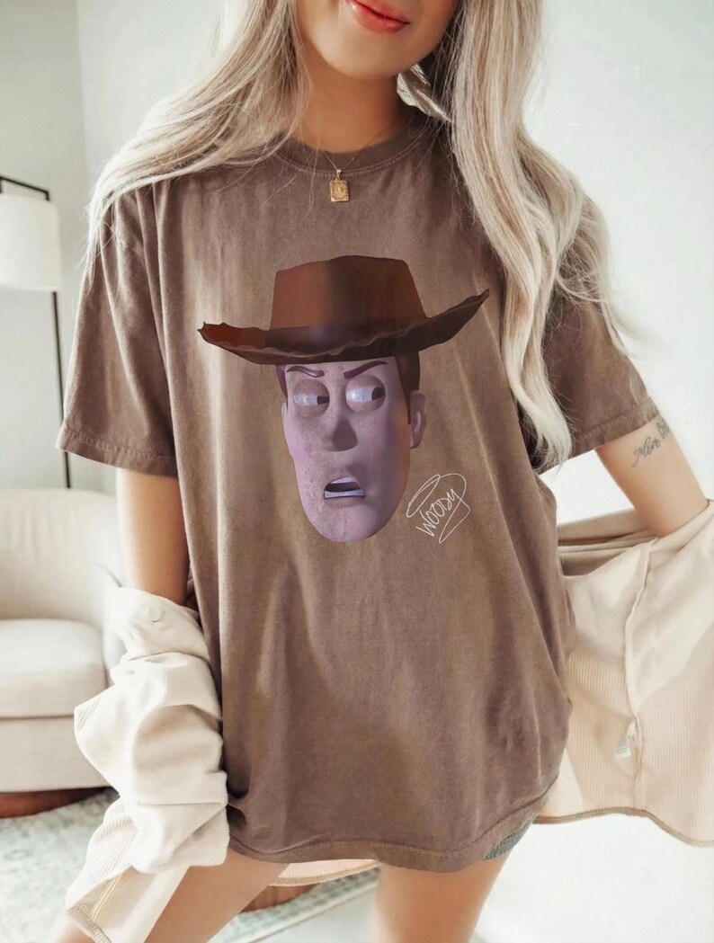 May include: A brown short-sleeved t-shirt featuring a graphic print of the character Woody from Toy Story wearing a brown cowboy hat. The character's face is centred on the chest, and the word Woody is written in a cursive script below the chin.