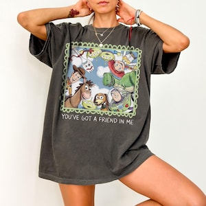 May include: Dark gray t-shirt featuring a colorful graphic of characters from the animated film Toy Story. The graphic is framed with a green scalloped border and includes the text "YOU'VE GOT A FRIEND IN ME".