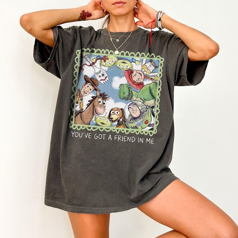 May include: Dark gray t-shirt featuring a colorful graphic of characters from the animated film Toy Story. The graphic is framed with a green scalloped border and includes the text "YOU'VE GOT A FRIEND IN ME".