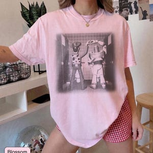 May include: A light pink short-sleeve t-shirt featuring a black and white graphic print of two cartoon characters standing at urinals in a tiled restroom, with one character forming a heart shape with their hands above their head.