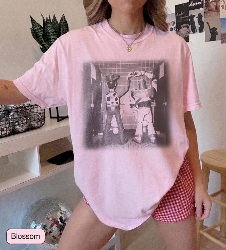 May include: A light pink short-sleeve t-shirt featuring a black and white graphic print of two cartoon characters standing at urinals in a tiled restroom, with one character forming a heart shape with their hands above their head.
