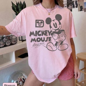 May include: A light pink t-shirt featuring a distressed graphic of Mickey Mouse. The design includes the words "Mickey Mouse" and a stylized image of Mickey. The shirt is short-sleeved and has a crew neck.