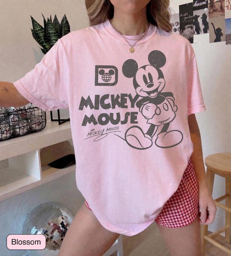 May include: A light pink t-shirt featuring a distressed graphic of Mickey Mouse. The design includes the words "Mickey Mouse" and a stylized image of Mickey. The shirt is short-sleeved and has a crew neck.