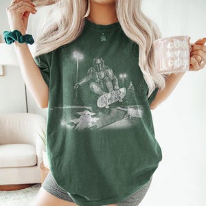 May include: A dark green short-sleeve t-shirt featuring a white graphic print of a character in armor skateboarding in a park at night, with a small creature standing nearby. The shirt has a crew neckline and a relaxed, loose-fitting silhouette.