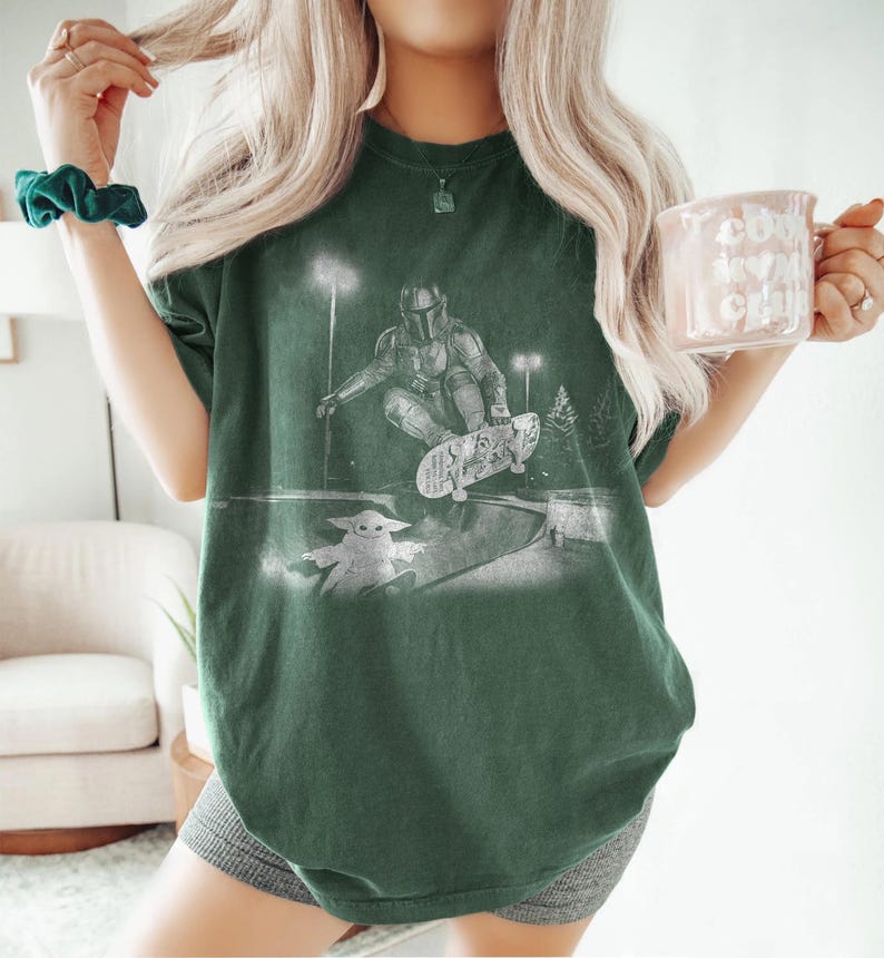 May include: A dark green short-sleeve t-shirt featuring a white graphic print of a character in armor skateboarding in a park at night, with a small creature standing nearby. The shirt has a crew neckline and a relaxed, loose-fitting silhouette.